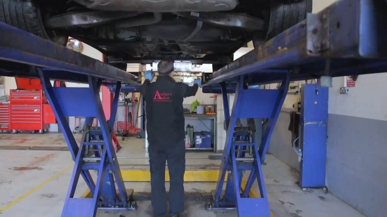 Mechanic walking beneath a car on a ramp at Archers Autos, from their promotional film