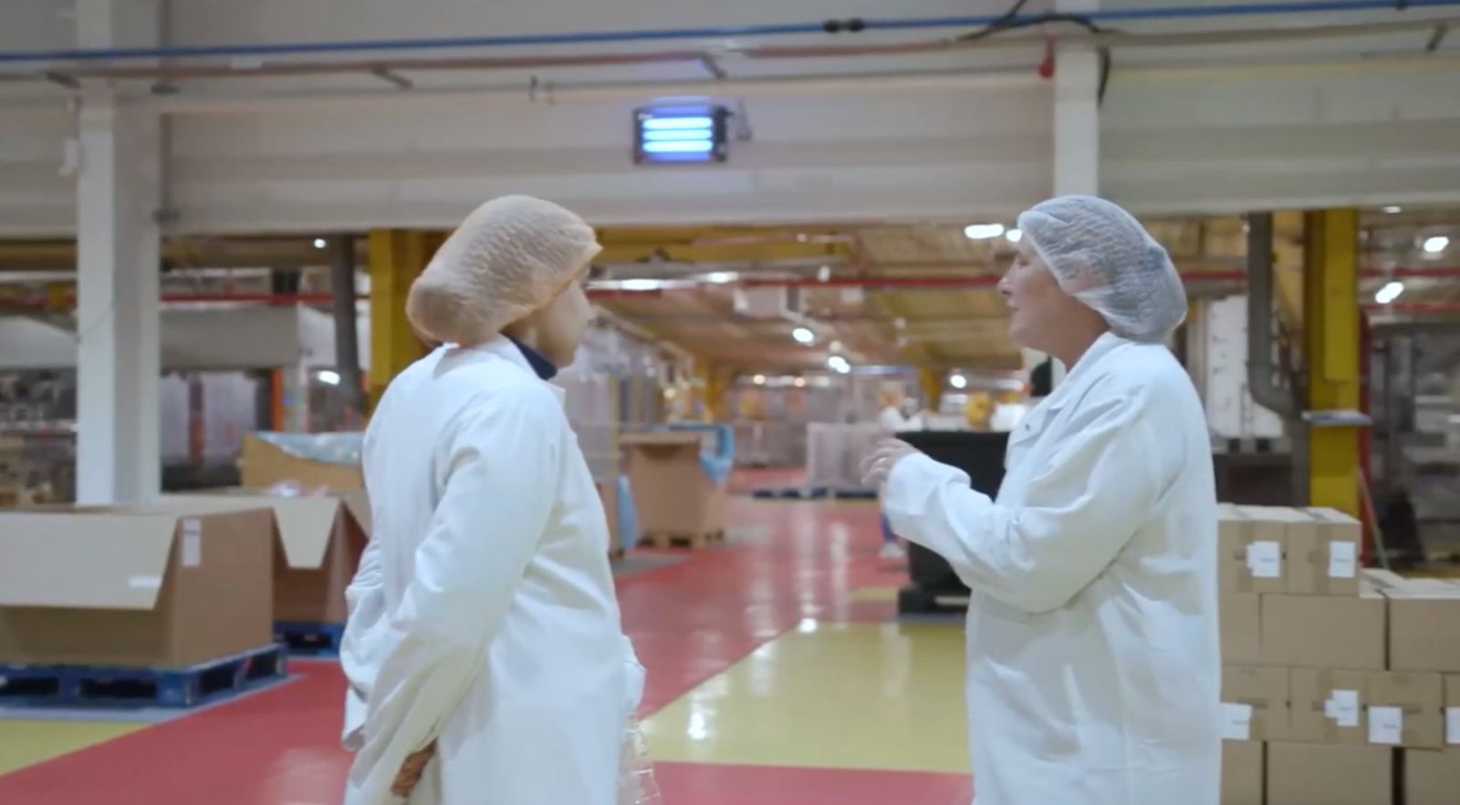 Two Magna staff in white coats and hairnets talking on the factory floor