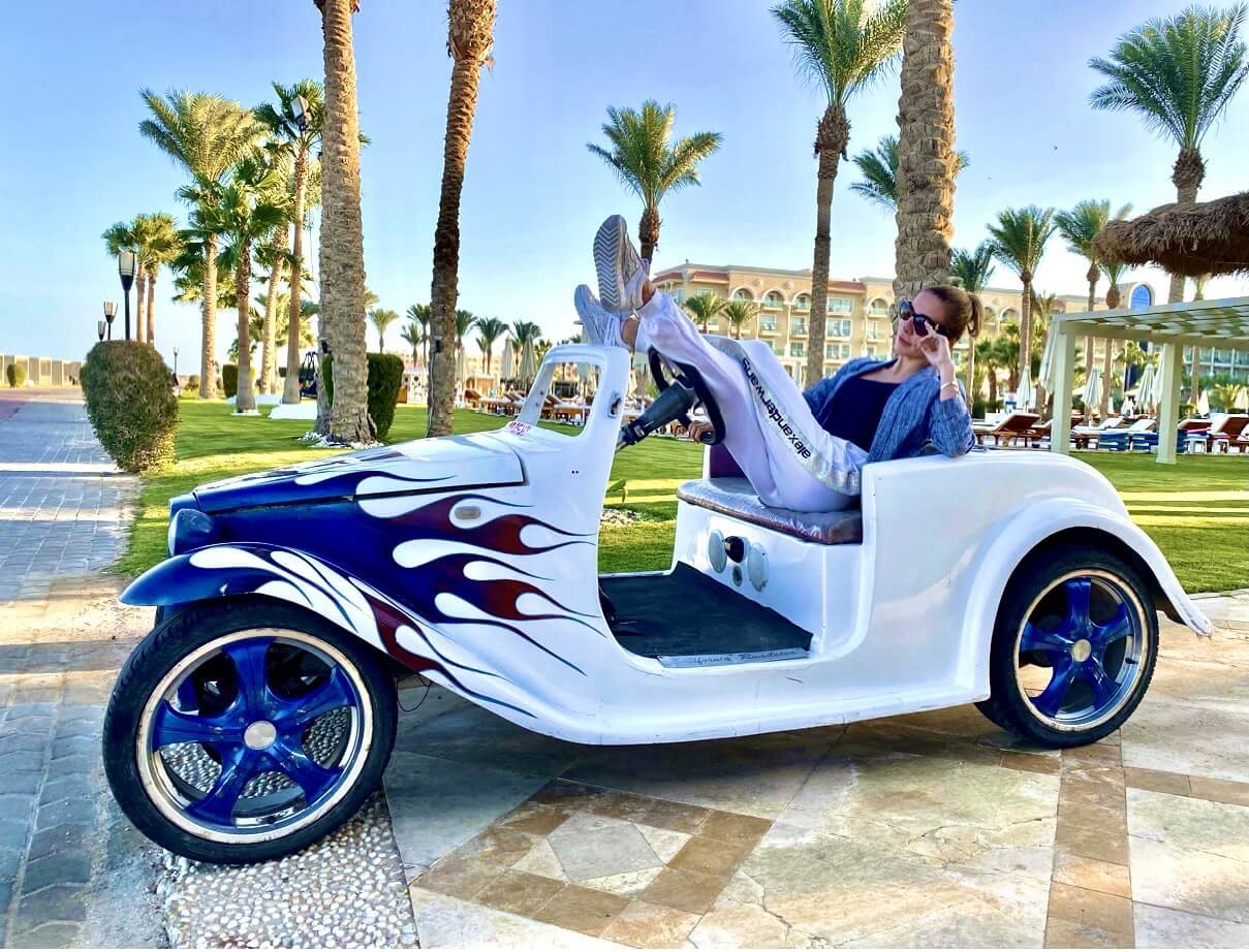 Marie in a three-wheeled sports car under palm trees
